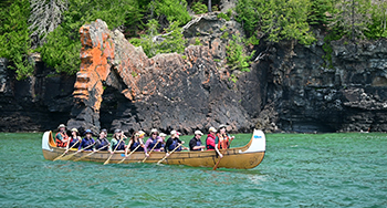 Un groupe de personnes portant des gilets de sauvetage pagaient dans un grand canoë sur une étendue d'eau bleue, avec une falaise rocheuse et une végétation luxuriante en arrière-plan.