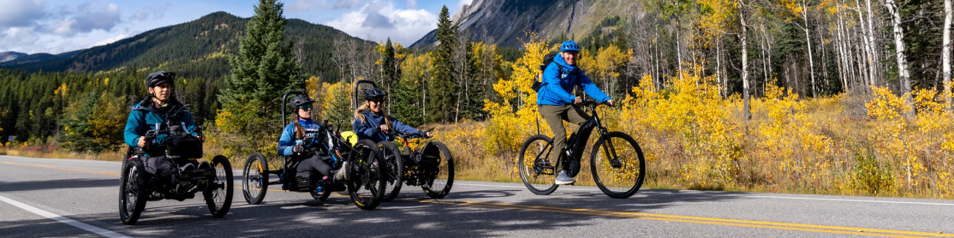 Un groupe de quatre cyclistes roule sur une route goudronnée en montagne par une journée ensoleillée. Trois cyclistes utilisent des handbikes adaptés, tandis qu’une personne à droite roule sur un vélo traditionnel. Ils sont entourés de hauts sapins et de feuillages d’automne jaunes vifs, avec des montagnes escarpées en arrière-plan.
