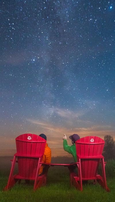 Two people sit in Parks Canada's red Muskoka chairs with their backs to the camera, looking up at a starry sky with the Northern Lights slightly visible.