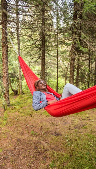 Une jeune femme noire allongée dans un hamac rouge attaché à un arbre dans une forêt d’arbres à feuillage persistant. Elle sourit.