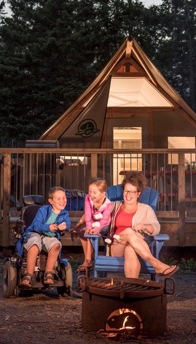 Une femme est assise sur une chaise muskoka bleue avec deux enfants à côté d’elle, ils rient. L’un des enfants est perché sur le bras de la chaise muskoka. L’autre est assis dans un fauteuil roulant électrique. Devant le groupe se trouve un foyer en métal sur lequel est découpé le logo de castor de Parcs Canada. Derrière eux se trouve une structure de tente semi-permanente (Otentik) avec un cadre en bois et un porche. La tente Otentik porte le logo du castor de Parcs Canada sur un rabat et est éclairée à l’intérieur.