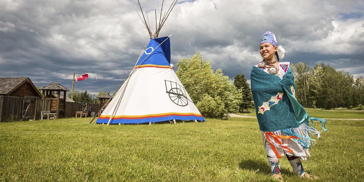 Indigenous girl wearing O'Chiese First Nation Junior Princess regalia in front of a tipi at Rocky Mountain House National Historic Site.