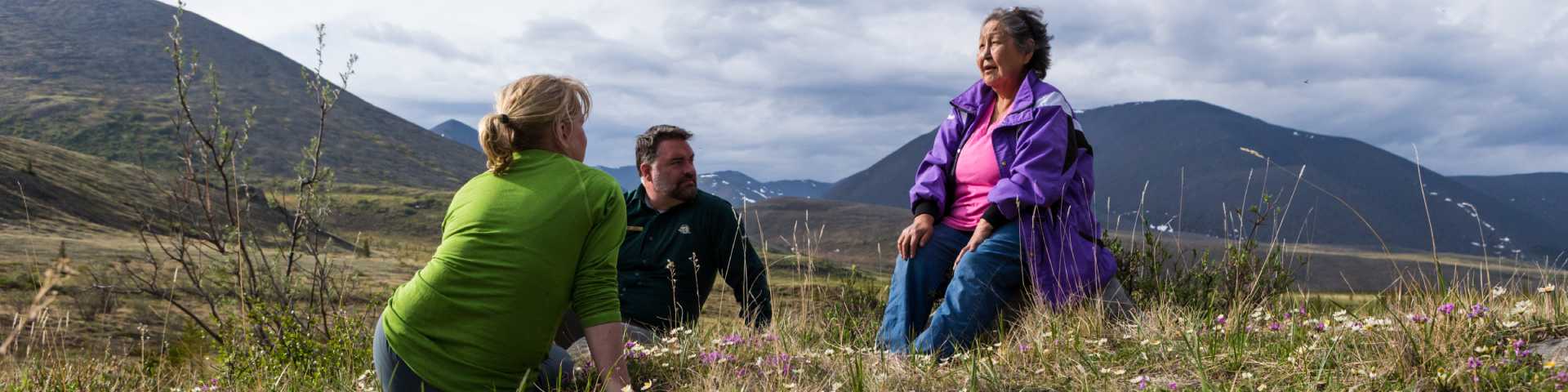 An indigenous elder sits with two Parks Canada employees in a mountain setting.