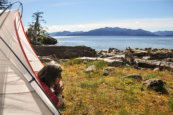 A woman laying down looks out of her tent over a beach and waterway