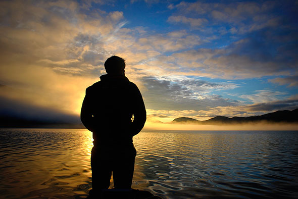 A man stands in front of a sunrise, looking over the calm water as mist burns off
