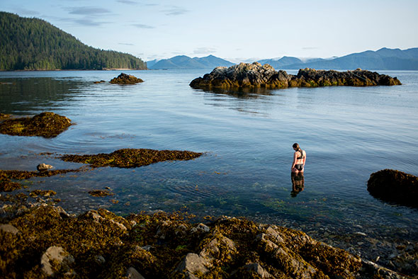 A woman stands knee deep in the water on a rocky coastline