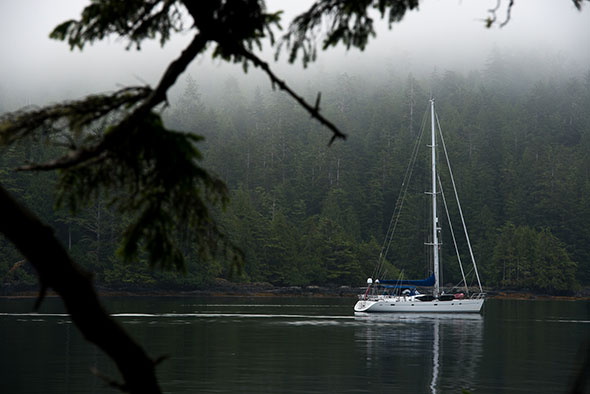 A sailboat motors on calm water under a low cloud layer