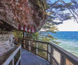 Wooden boardwalk along a rocky shoreline, overlooking blue water and trees.