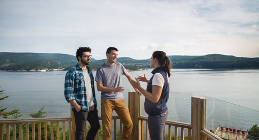 Deux visiteurs discutent avec une guide-interprète au centre d'interprétation et d'observation de Pointe-Noire. Derrière eux se trouve l'embouchure de la rivière-Saguenay. 