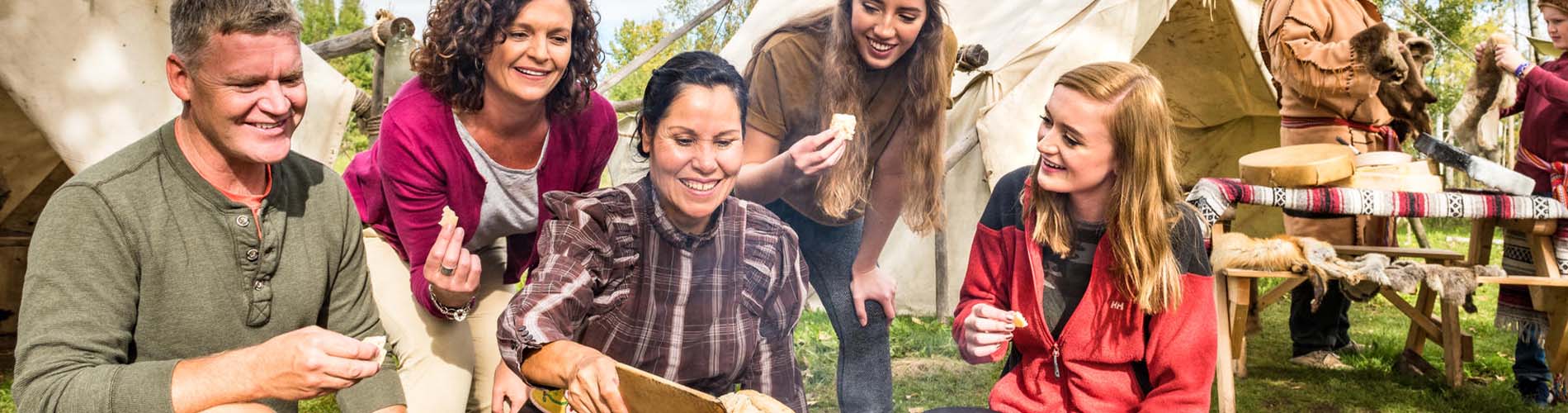 An Indigenous woman animator cooks up bannock to offer a family of visitors at the Métis Campfire. A boy with Métis animator discuss pelts and handmade drums in the background. Rocky Mountain House National Historic Site.