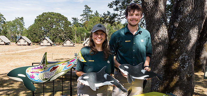 Photo of two Parks Canada interpreters in uniform in front of a table showing killer whale information. 