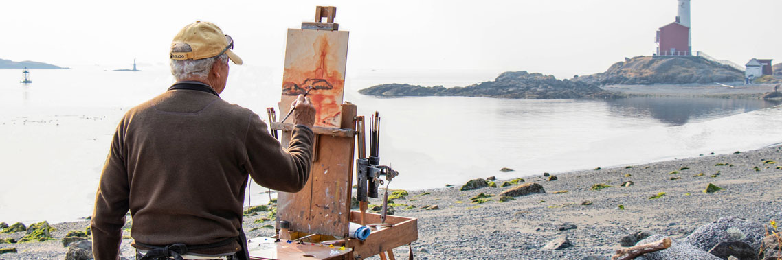 On the beach, a person uses an easel to create a painting of Fisgard Lighthouse.
