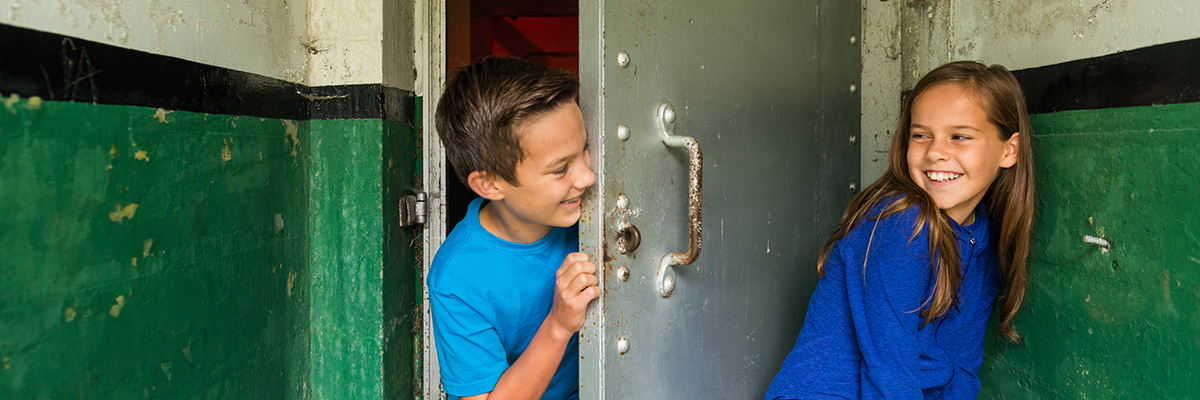 Two children play hide-and-sike; one of the children is hiding behind a steel door with rivets.