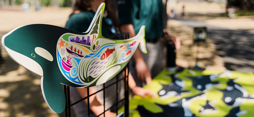 A cardboard whale with fish, waves, boats, a city and other marine animals drawn inside, on a table lined with whales.