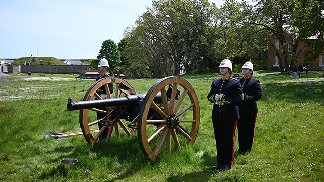 Three Parks Canada employees in uniform stand beside a large historic cannon on a grassy field under a clear sky.