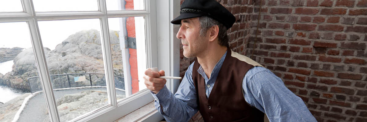 An interpreter in a lighthouse keeper costume looks a pipe in hand out the window.