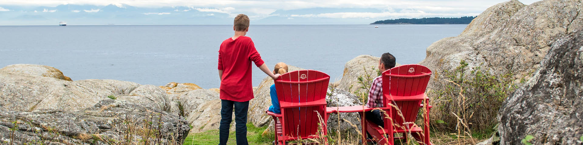 A couple sitting on red chairs with their child standing beside them, all three are admiring the view of the ocean and mountains.