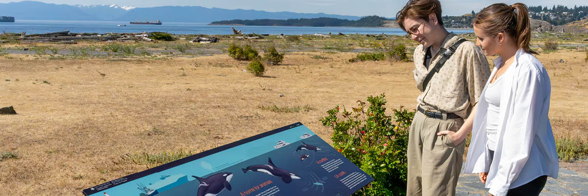 A couple stand in front of a whale sign in front of the ocean with a passing freighter and mountains in the background.