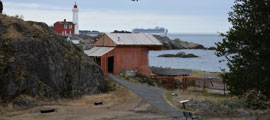 A gravel path leads down to a searchlight emplacement disguised as a red fisherman’s hut. Beyond it is a rocky beach. The lighthouse is visible in the background.