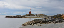 In the foreground is a pebbled beach with a gentle slope. There is a seaweed covered rocky outcrop on the right hand side. In the background is the lighthouse and causeway.