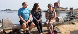 A couple of seniors with their adult daughter sit on a wooden bench, with Fisgard Lighthouse in the background.