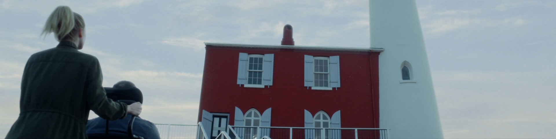 Au pied du phare de Fisgard, une personne en fauteuil roulant et son accompagnatrice admirent le phare.
