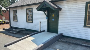 Bâtiment en bois blanc, doté de fenêtres aux encadrements verts et d'une porte verte, surmonté d'un petit toit et muni d'une rampe d'accès en béton pour fauteuils roulants.