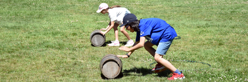 Two children outdoors rolling wooden barrels on a grassy field during a sunny day.
