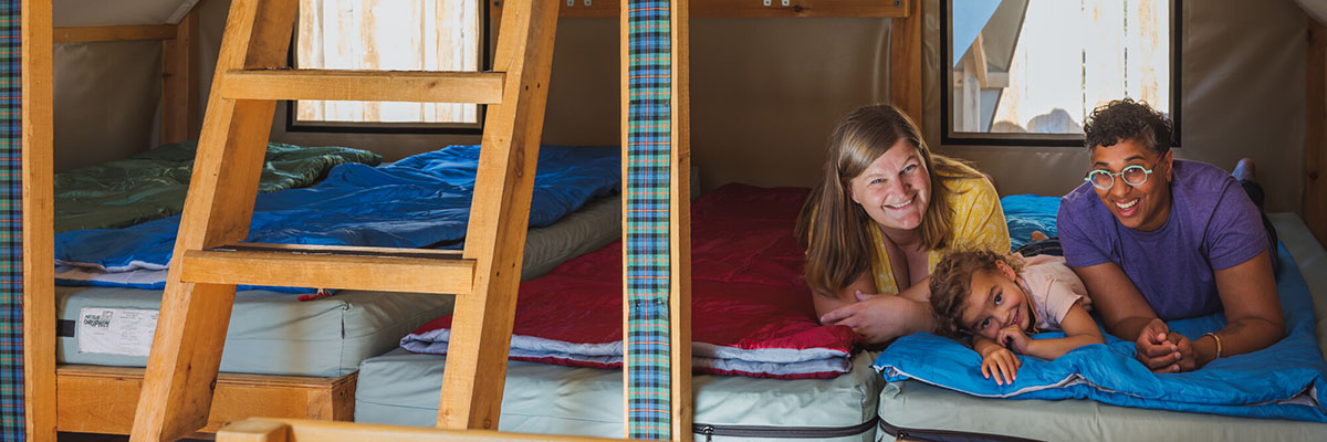 Deux parents allongés avec leur enfant entre eux sur l’un des lits de la tente oTentik, souriant tous ensemble.