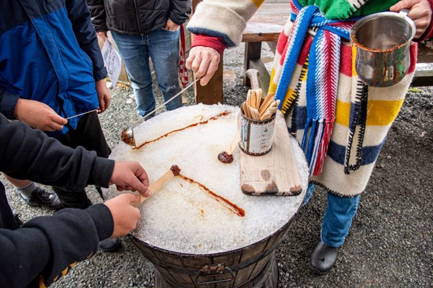 A person in historical costume pours maple syrup onto snow while a visitor rolls a wooden stick in the hardened syrup.