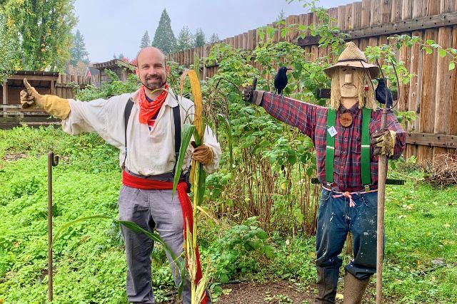 Une personne en costume du XIXe siècle posant et souriant à côté d’un épouvantail dans un jardin.