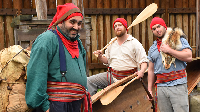 A group of historical interpreters in period clothing posing with a canoe, paddles, and fur pelts.