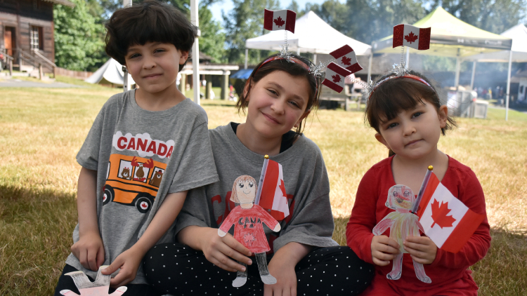 Three children outdoors wearing Canada-themed clothing and holding Canadian flags on a sunny day. 