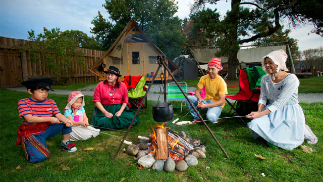 Group of people in historical clothing sitting around a campfire roasting food on sticks in a backyard setting.
