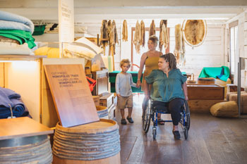  Deux adultes et un enfant visitent l’exposition présentée à l’Entrepôt. L’un des adultes se déplace en fauteuil roulant. Derrière eux, une collection de fourrures est accrochée au mur de l’exposition.