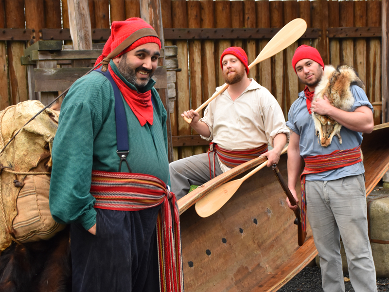 Trois interprètes costumés portant des écharpes Voyageur, des capots et tenant des pagaies en bois