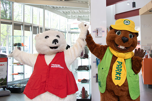 La mascotte d’ours polaire, vêtue d’un gilet rouge portant l’inscription « Travel Manitoba », tape dans les mains de la mascotte de castor, coiffée d’un chapeau jaune, vêtue d’un gilet vert et d’une cravate jaune portant l’inscription Parka