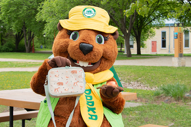 Une mascotte de castor tient un panier-repas dans une main, une pomme et une brindille dans l’autre.