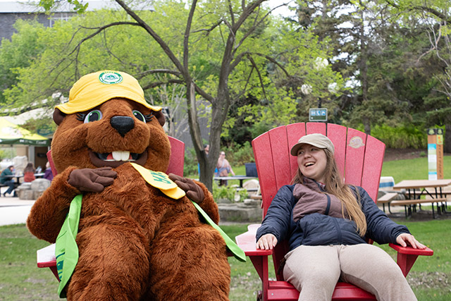 A beaver mascot leans back in a red Adirondack chair while a visitor also leans back in a chair and smiles at her.