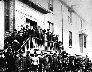 Cette photo en noir et blanc montre des personnes se tenant dans la rue et le long d’un escalier devant un bâtiment sur lequel sont peints, au-dessus de la porte, les mots « Dominion Government Immigration Hall ».