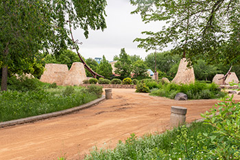 Green plants with yellow flowers are shown to the right of a wide brown path, and green plants with white flowers are shown to the left. Five beige brick structures can be seen in the background.