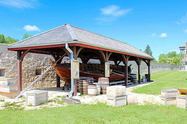 A York boat sits underneath a wooden structure with several barrels and crates nearby.