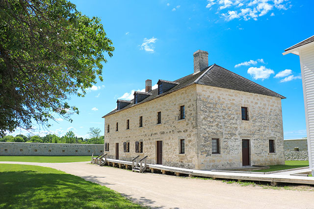 A gravel path and wooden boardwalk lead to a light-coloured three-story stone building.