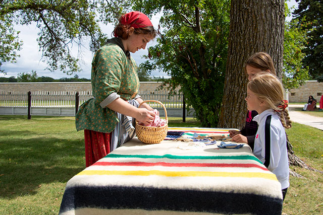 A Parks Canada employee in historic clothing gestures to fabrics on a table as two children watch at Lower Fort Garry.