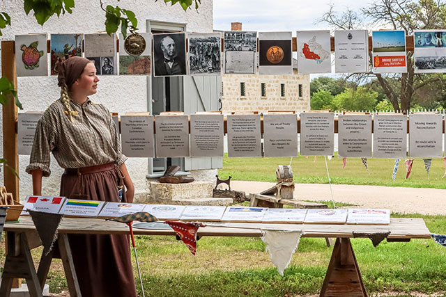 A Parks Canada employee in historic clothing stands in front of a display with one line of images in a row and a line of pages of text in a row below it.