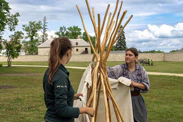 Two Parks Canada employees, one in uniform and one in historic clothing, assemble a miniature tipi.