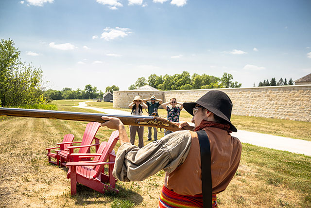 A Parks Canada employee in historic clothing aims a historic weapon as three people cover their ears in the background.
