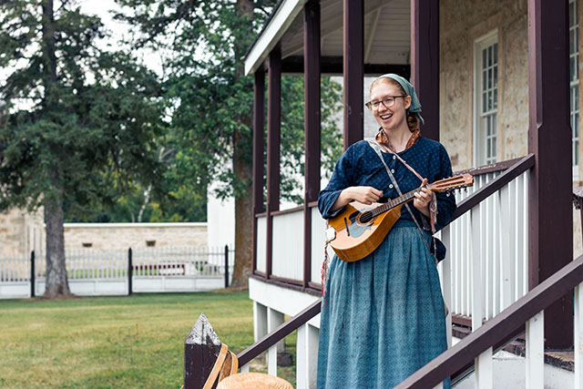 A Parks Canada employee in historic clothing strums a stringed instrument and smiles at Lower Fort Garry.