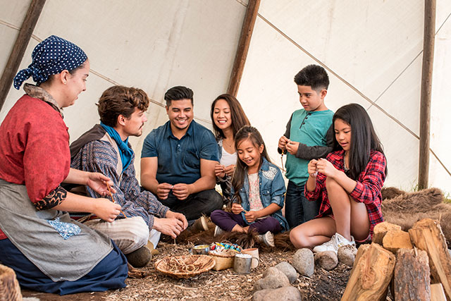 Two Parks Canada employees in historic clothing and five guests sit inside a tipi.