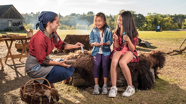 Deux enfants assis sur une balle de fourrure sourient tout en interagissant avec une employée de Parcs Canada en tenue d’époque.